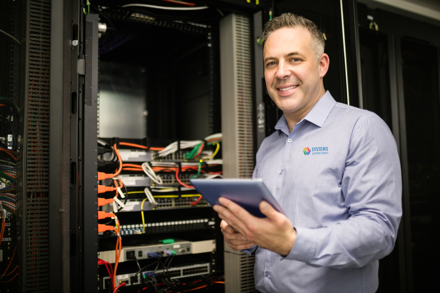 A man smiling with a tablet in hand in front of server racks.